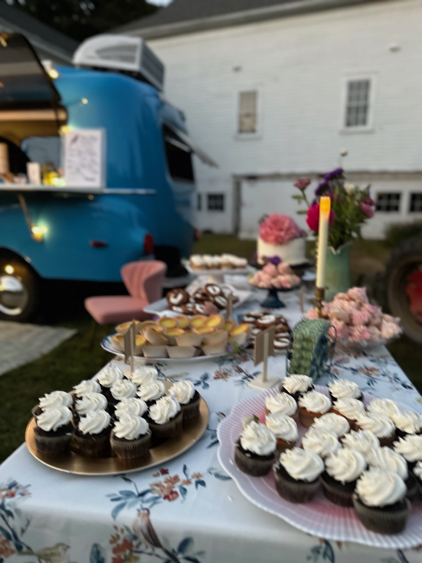 A curated dessert table with cupcakes, pretzels, and a wedding cake beside the Blue Bird camper at an evening outdoor event